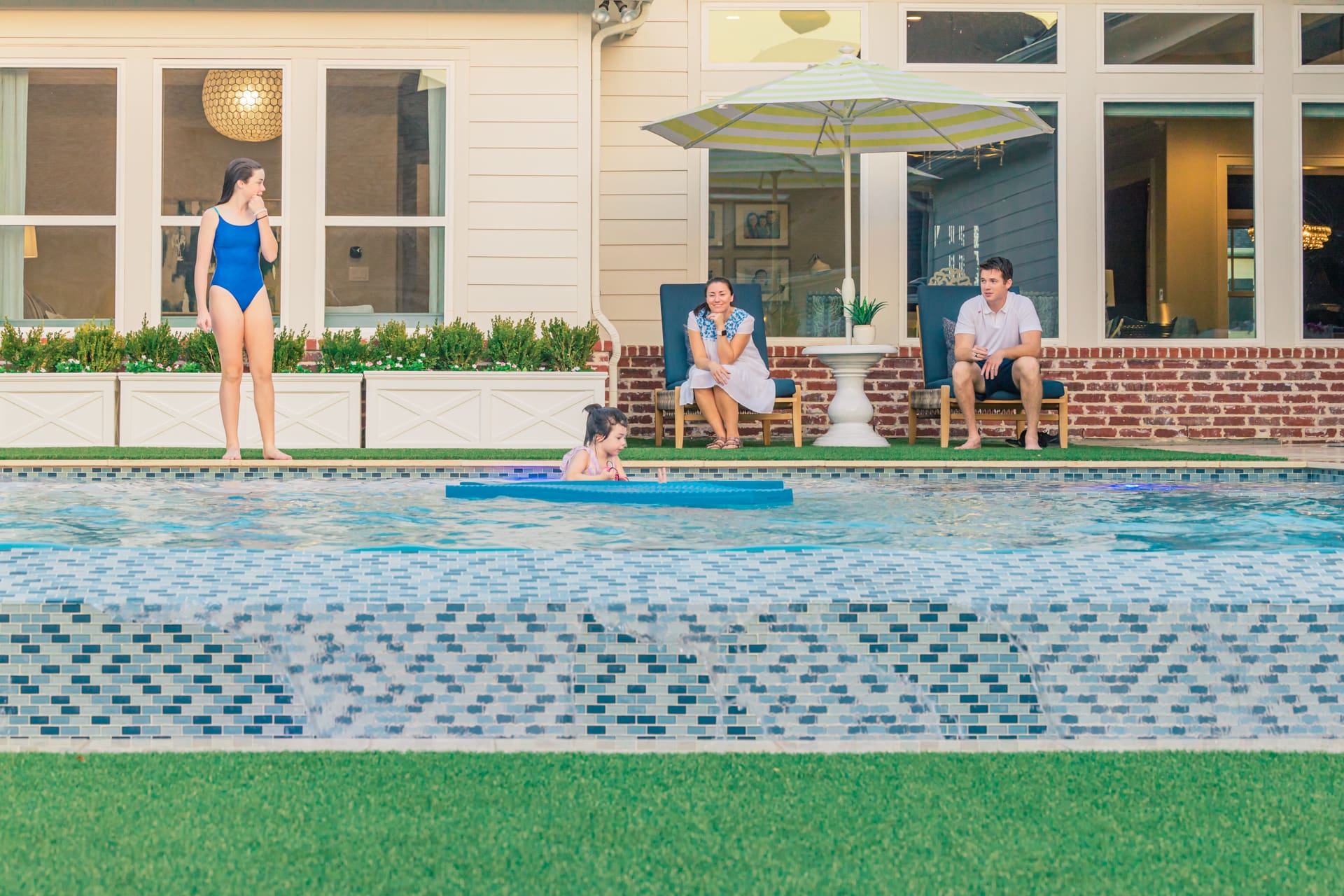 A young girl swims on a foam mat in a backyard pool. An adult woman in a blue swimsuit stands at the pool's edge, while two adults sit on patio chairs under an umbrella. The pool is bordered by a garden and a light-colored house with large windows.