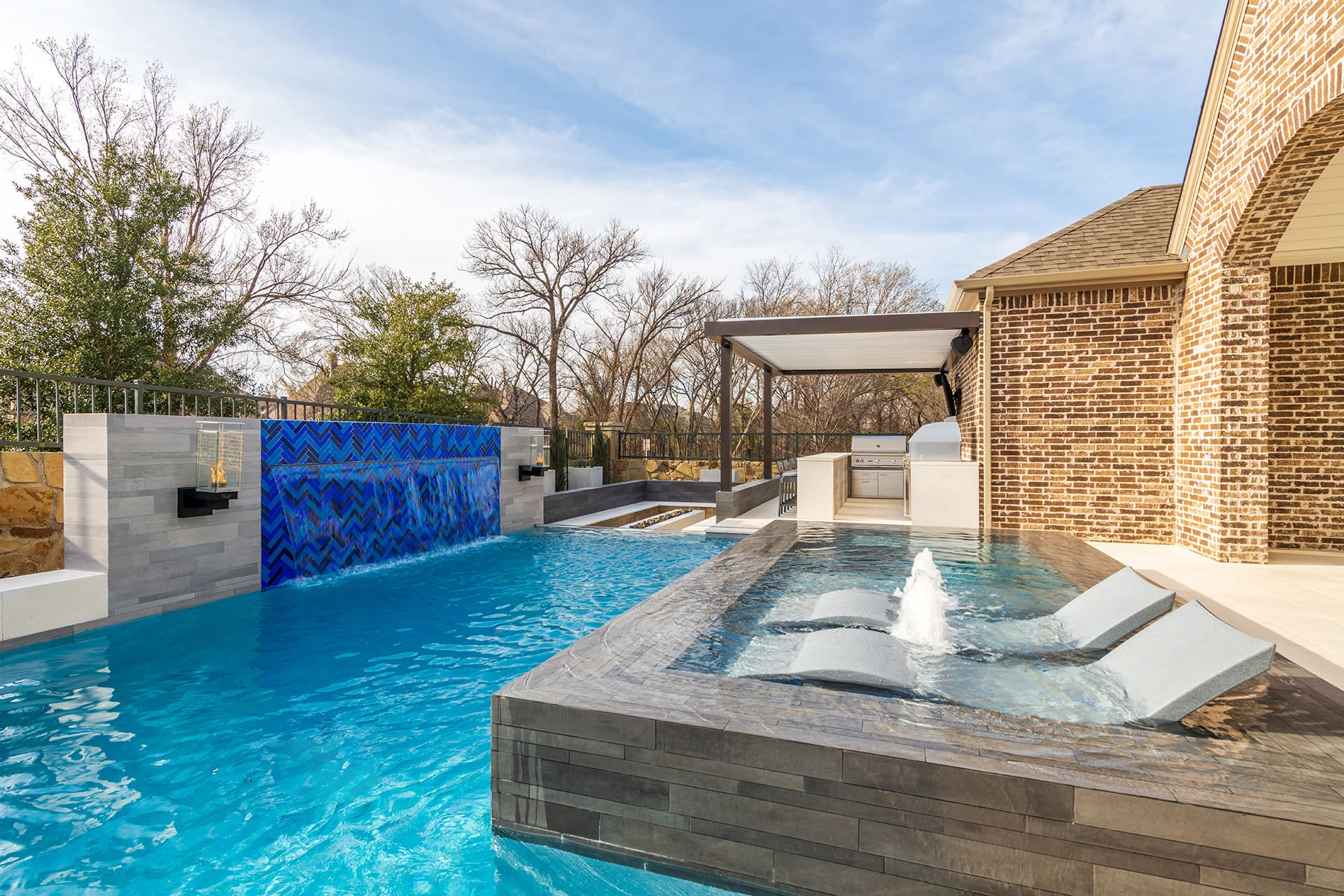 A modern outdoor swimming pool with built-in lounge chairs and a cascading waterfall feature. The pool area includes a shaded patio with a barbecue station and bar-style seating, surrounded by brick and stone structures. Trees and a blue sky are visible in the background.