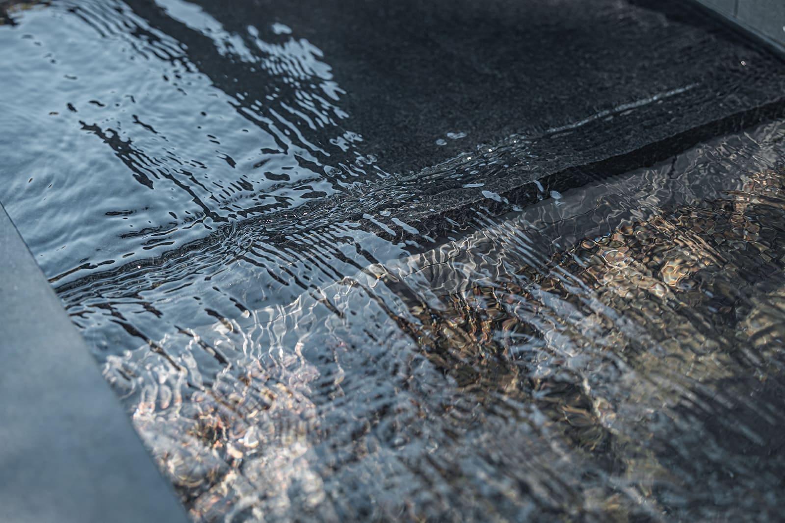 A close-up view of water gently rippling across a smooth, dark surface, reflecting light and creating intricate patterns. The water appears calm and clear, capturing a mix of light and shadows, with a serene and soothing ambiance.