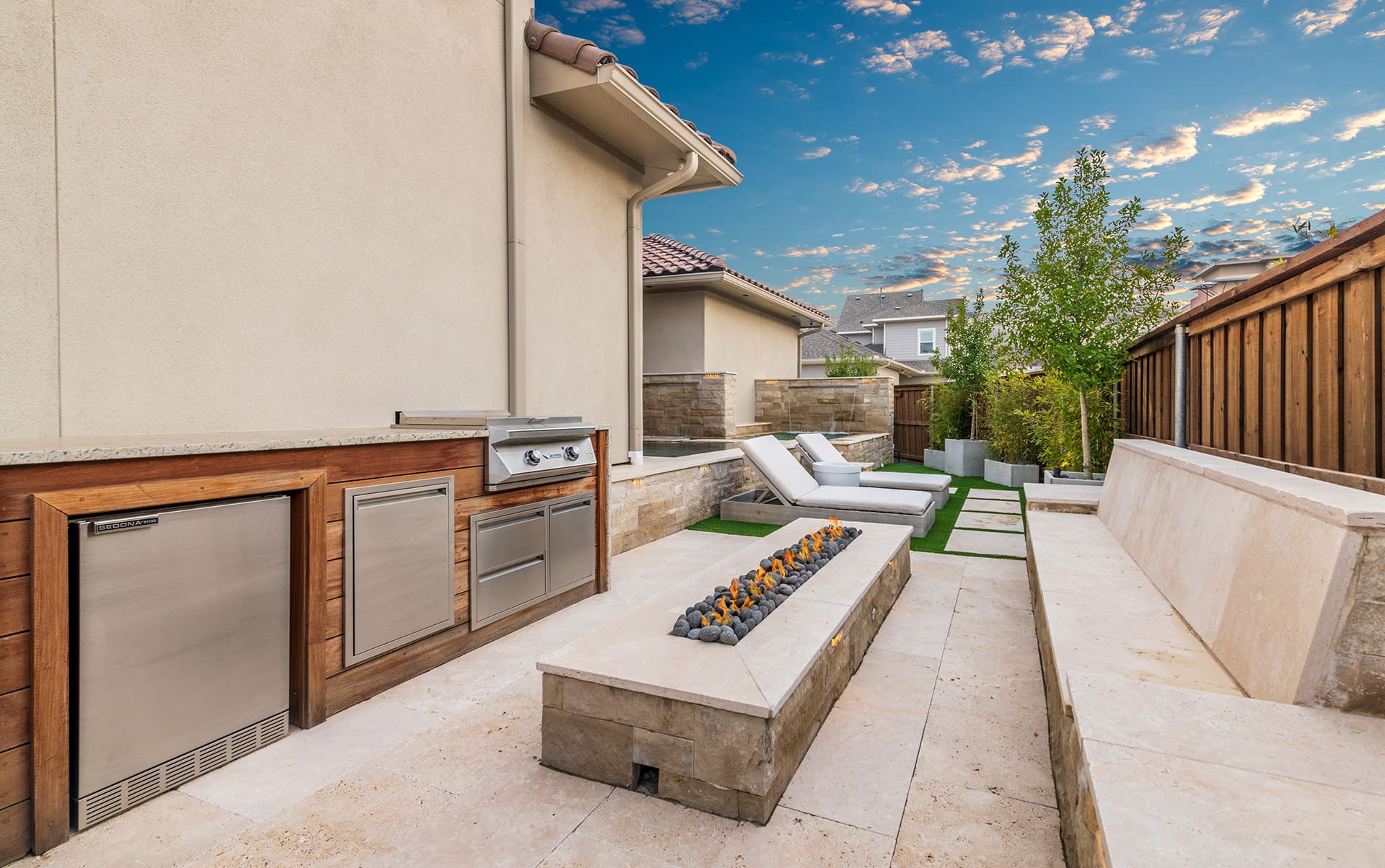 A modern backyard patio with an outdoor kitchen featuring a stainless steel grill and refrigerator. There’s a stone fire pit in the center, surrounded by cushioned lounge chairs. The area is enclosed by a wooden fence and greenery, with houses visible in the background.