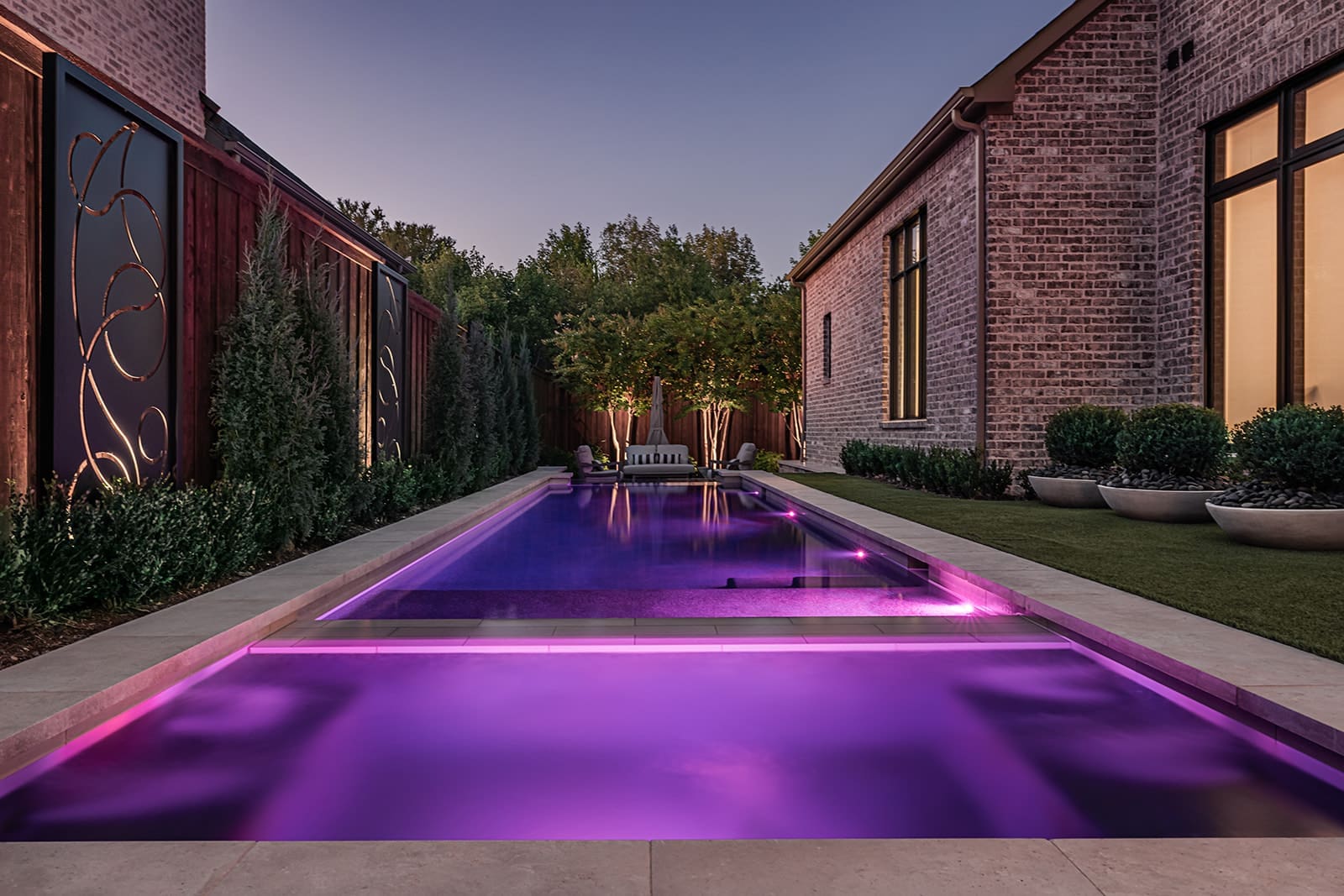 A luxurious backyard pool illuminated with purple lighting at dusk, surrounded by manicured plants and modern decor. The pool is flanked by two brick buildings with large windows, and there's a serene, well-lit seating area with trees in the background.
