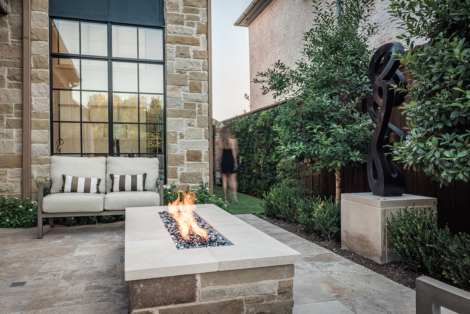 A cozy outdoor patio features a stone fireplace with a lit fire, surrounded by lush greenery and a modern art sculpture on a pedestal. A cream-colored sofa with striped cushions sits close by, and a person in black is seen walking in the background.