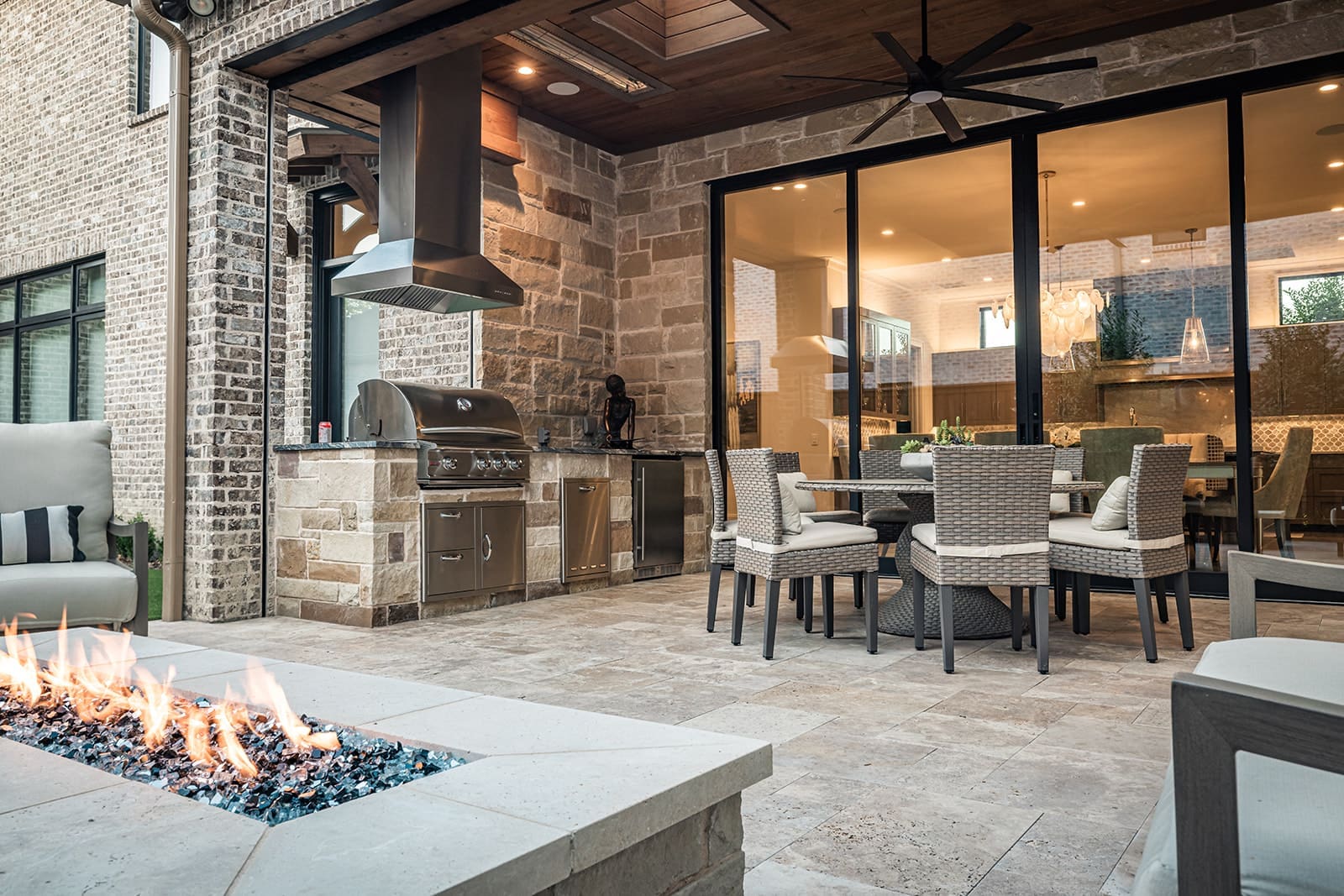 Outdoor patio with modern kitchen featuring stone walls, stainless steel grill, range hood, and dining area with wicker chairs and table. Glass doors lead into the house. Fire pit with glass rocks in the foreground, set on tiled flooring. Ceiling fan above.