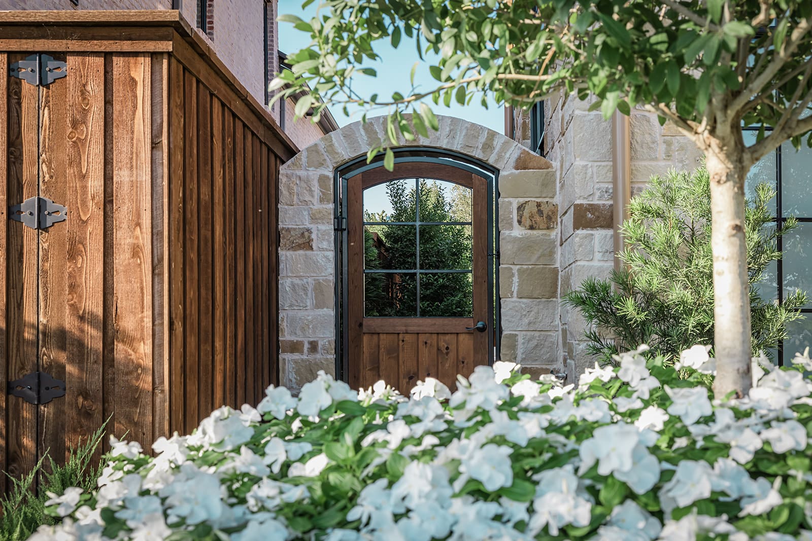 A charming garden scene featuring a wooden and stone archway leading to a wooden gate with glass windows. Lush green shrubs and white flowers bloom in the foreground, and a tree provides partial shade.