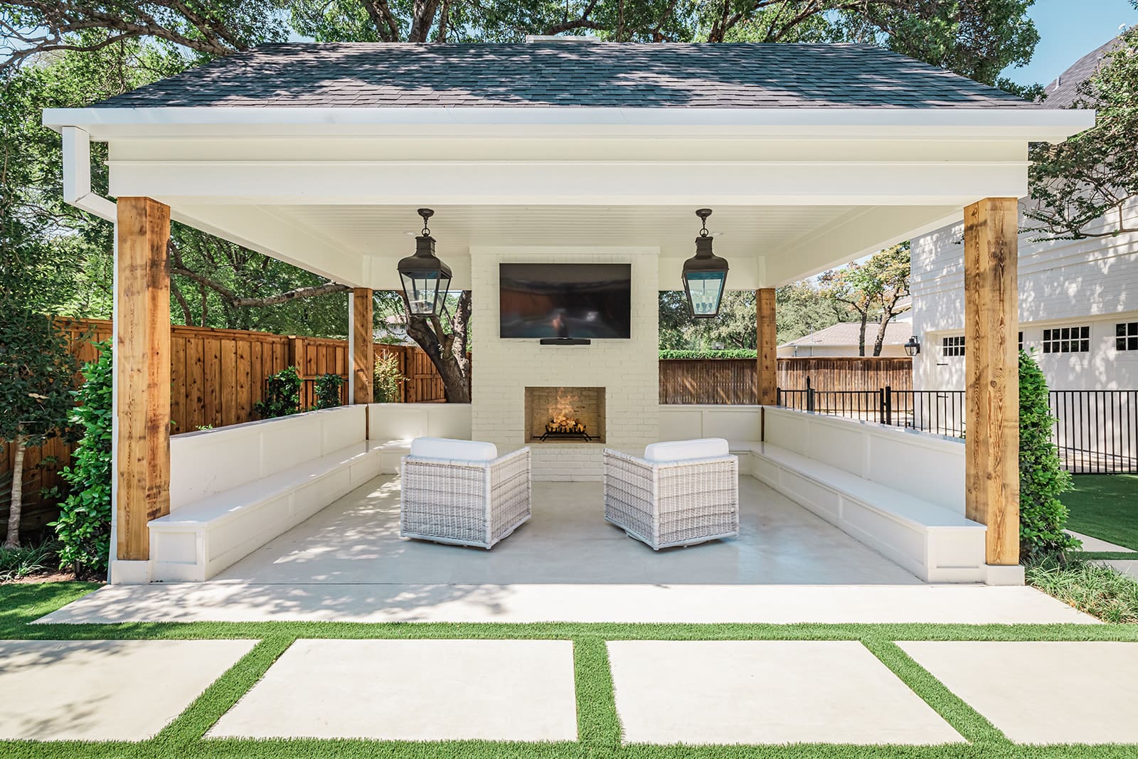 A modern outdoor pavilion features white cushioned seating along three sides, a central fireplace, and a mounted TV above it. Two lantern-style lights hang from the ceiling. Two wicker chairs face each other at the center, with a stepping stone path leading to the pavilion.