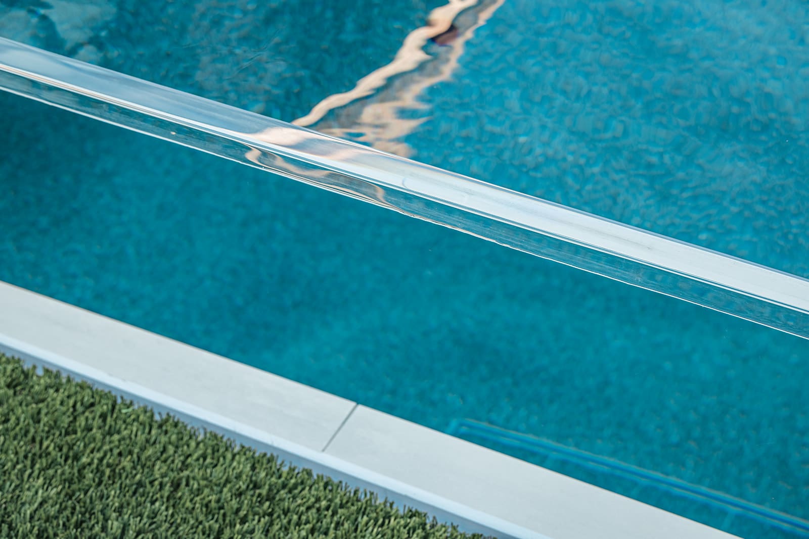 A close-up view of a modern swimming pool with clear blue water. The pool is bordered by a low, transparent glass barrier and a strip of green artificial turf. The water surface has visible reflections creating gentle ripples.