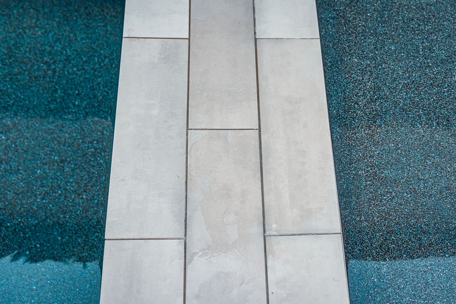 A top-down view of a partially submerged rectangular concrete slab surrounded by deep blue pebbles. The slab has a few droplets of water on its surface, suggesting it is adjacent to a watery area, possibly a pool or water feature.