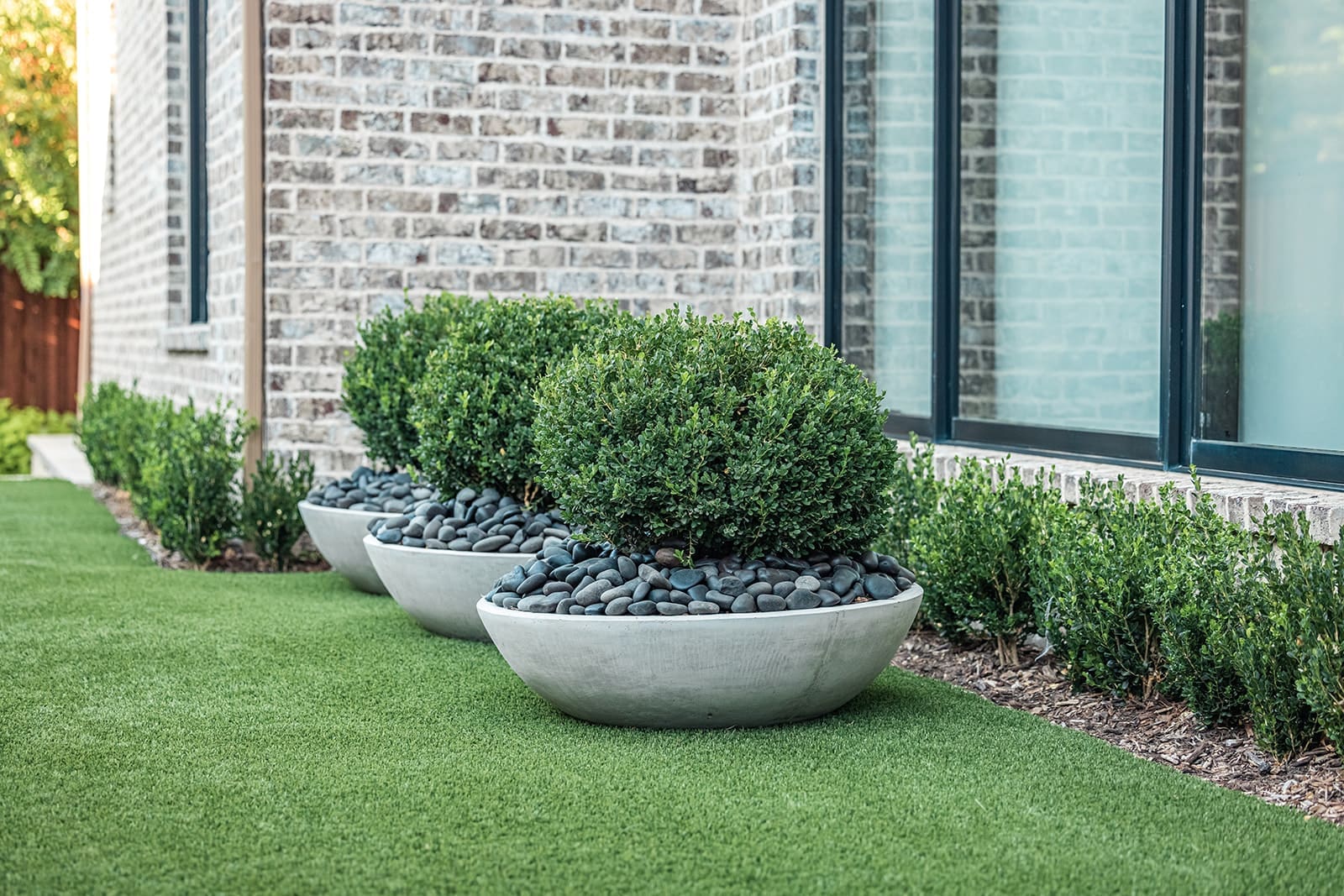 Three large, round concrete planters filled with smooth black stones and trimmed hedges are placed alongside the corner of a brick building. The planters are set on neatly manicured artificial grass, with additional small shrubs planted along the building's foundation.