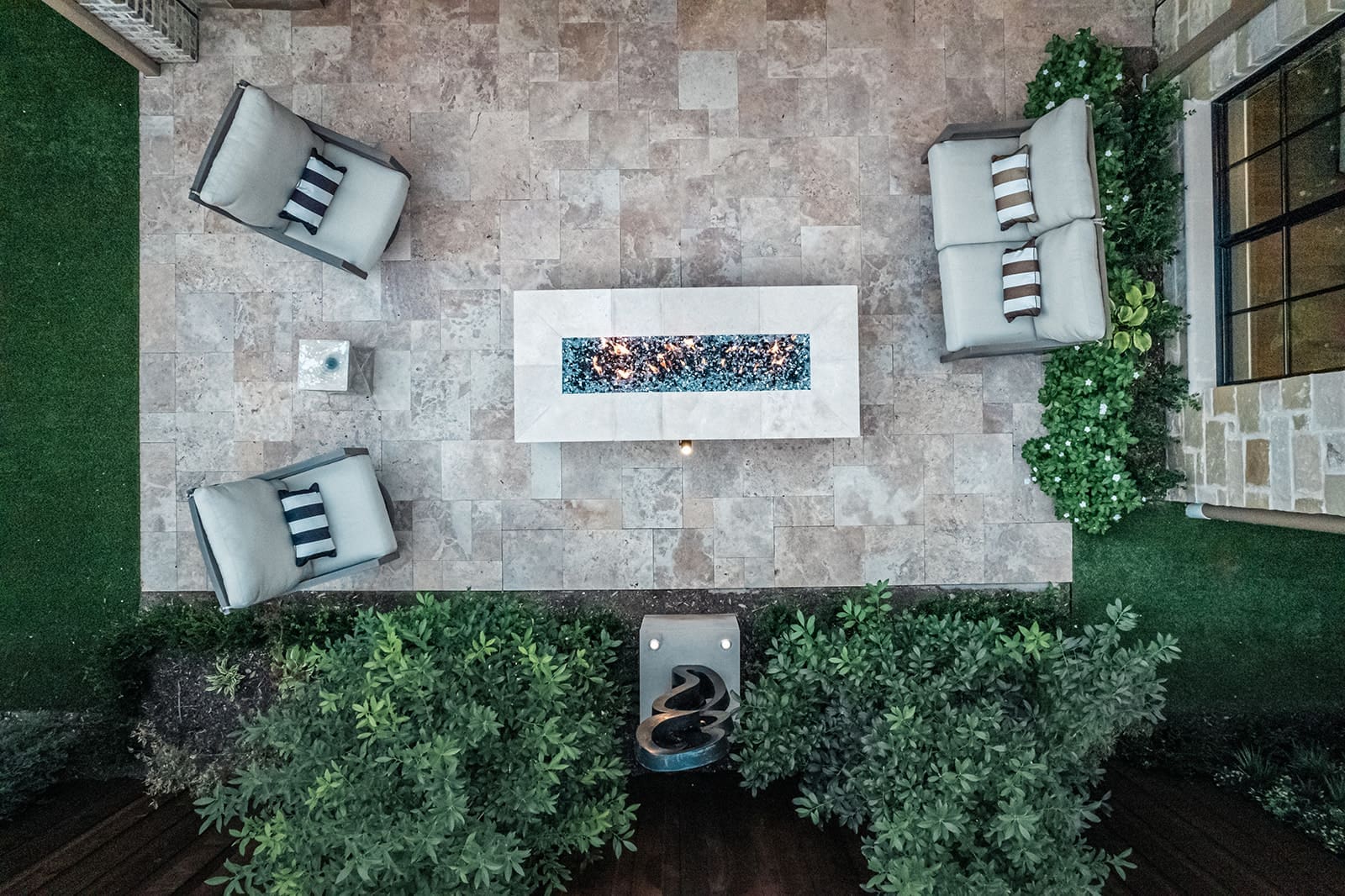 Aerial view of an outdoor patio with a rectangular fire pit in the center. Four cushioned chairs with striped pillows are arranged around the fire pit. The patio is bordered by greenery, and there's a modern sculpture near one end. The floor is tiled in neutral tones.
