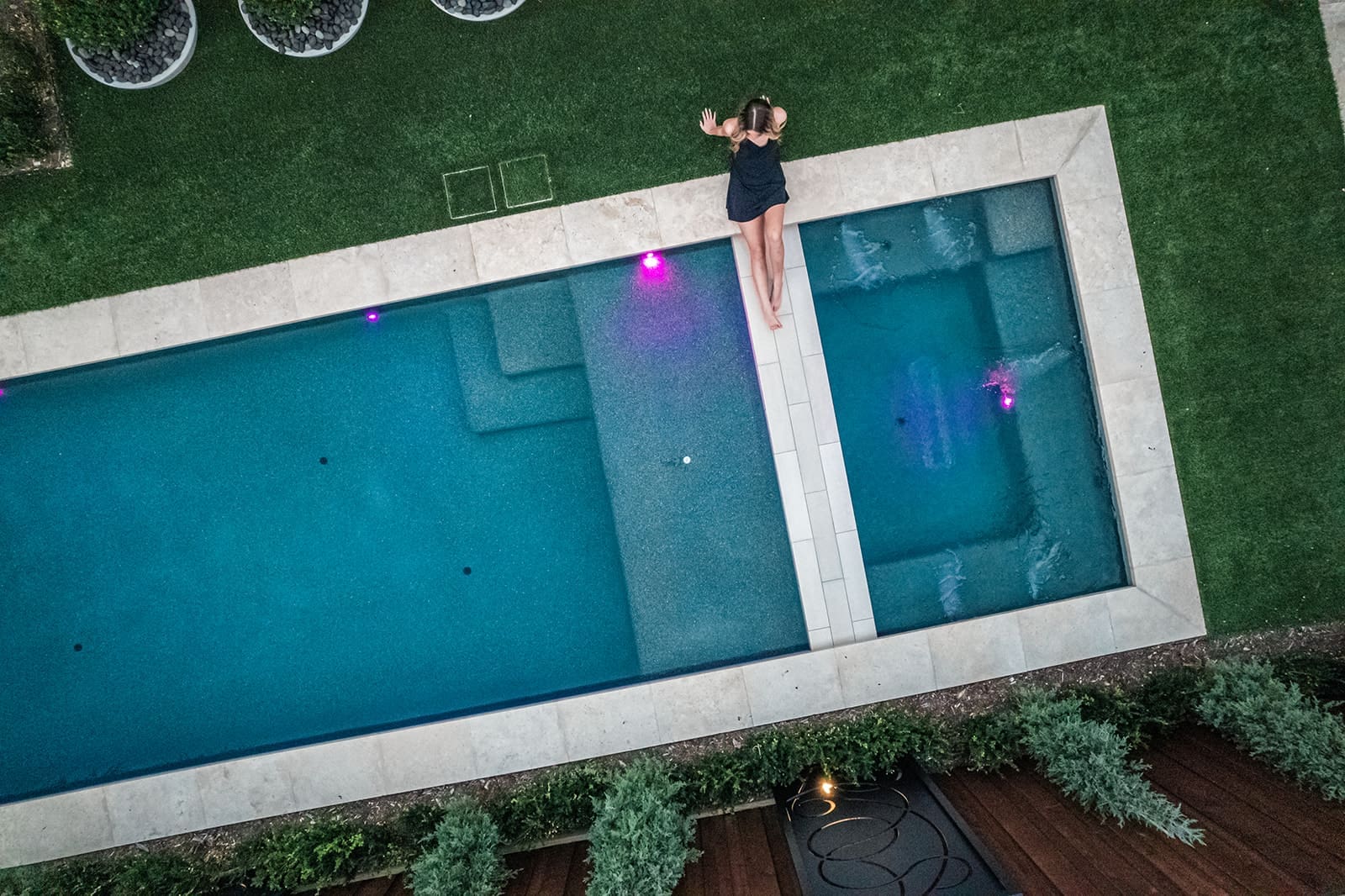 Aerial view of a person in a black outfit lying on the edge of a rectangular swimming pool with their legs in the water. The pool is surrounded by green grass and has purple lights illuminating the water.