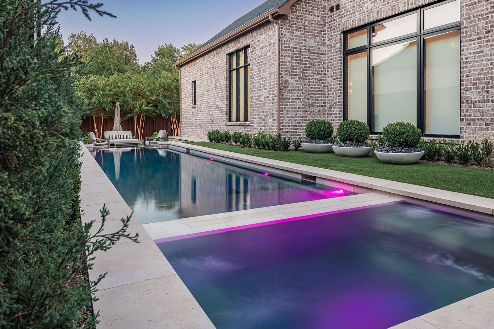 A modern backyard features a sleek rectangular pool with purple lights under the water. It is surrounded by a stone patio and well-manicured green shrubs. Adjacent to the pool is a brick house with large windows. In the background, there are lounge chairs and trees.