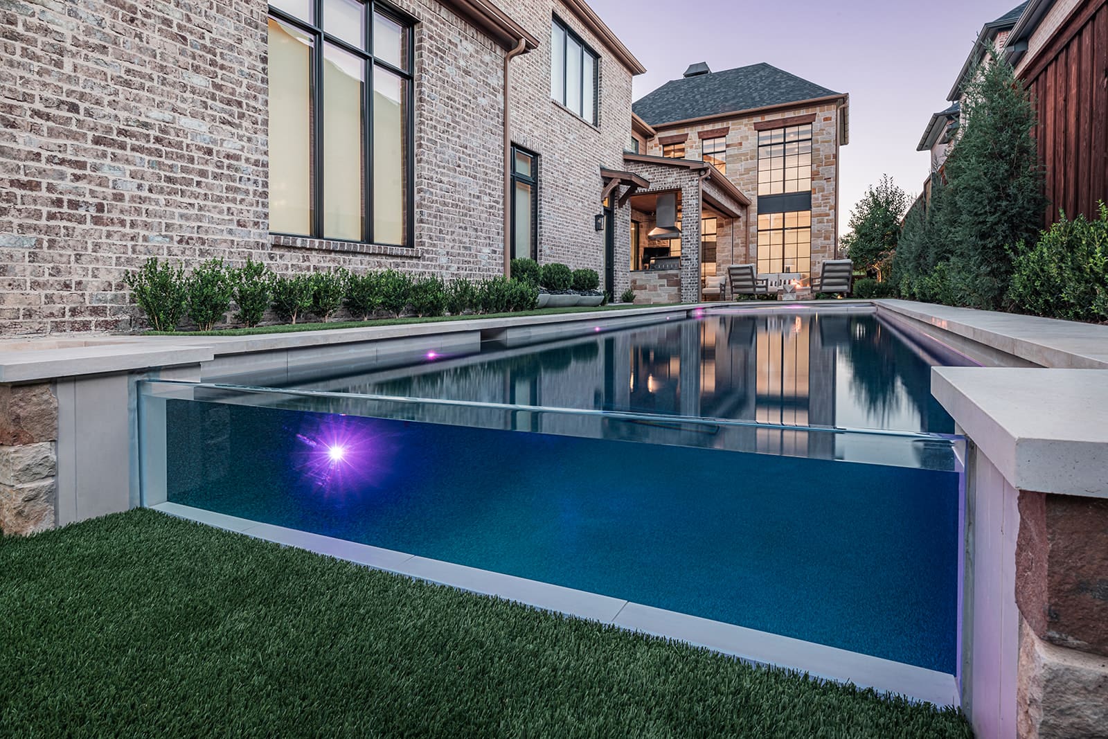 A modern backyard features a long rectangular pool with a transparent glass end, revealing the water inside. The pool is surrounded by lush green grass, brick houses, and manicured shrubs. Lounge chairs and an outdoor shaded seating area are seen in the background.