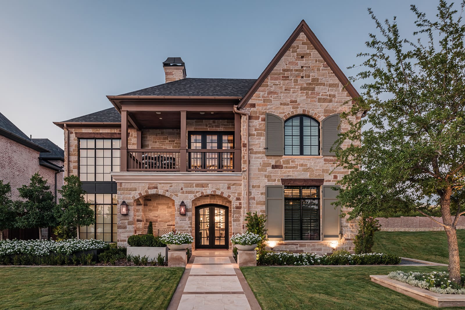 A two-story stone house with large windows, a covered balcony, and a neatly manicured lawn. The front entrance has two lantern-style lights and a paved walkway. The home features a mix of stone and siding, with greenery and flowers lining the walkway.