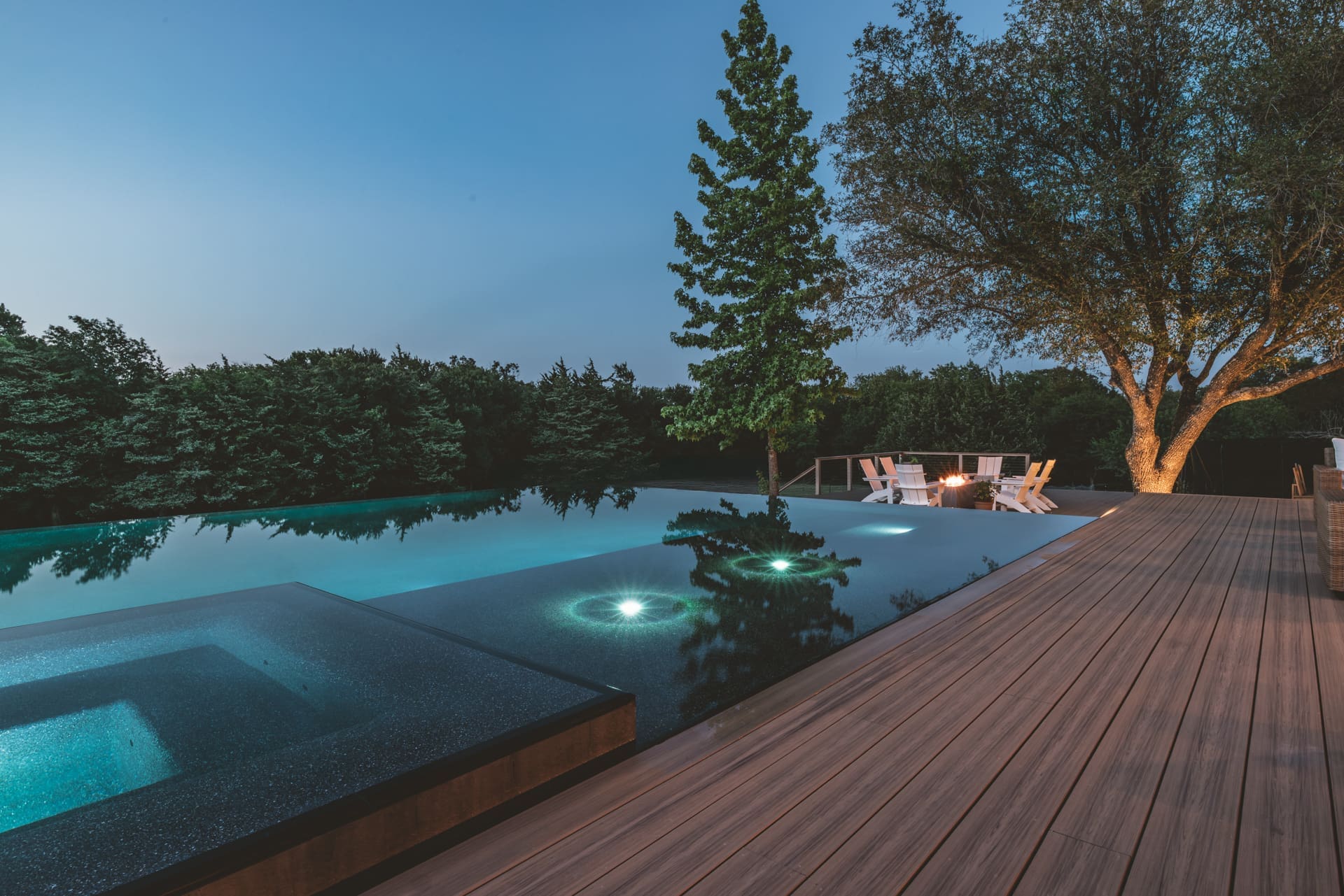A serene backyard scene at dusk featuring an infinity pool with glowing underwater lights. Surrounding the pool are trees, and a wooden deck extends along the edge. A cozy fire pit with chairs is visible under a large tree.
