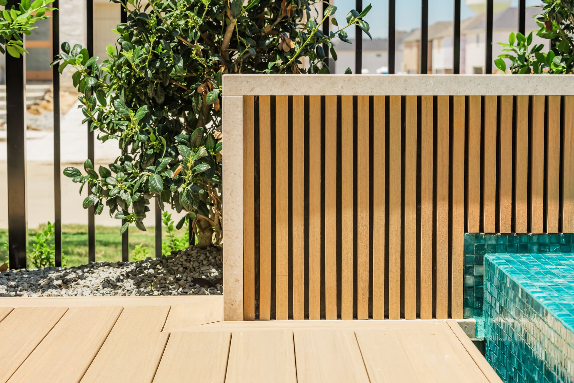 A modern outdoor space featuring a light wooden deck and vertical wooden slats adjacent to a small tiled pool. Green foliage and a black metal fence line the background, with a sunny, clear sky illuminating the scene.