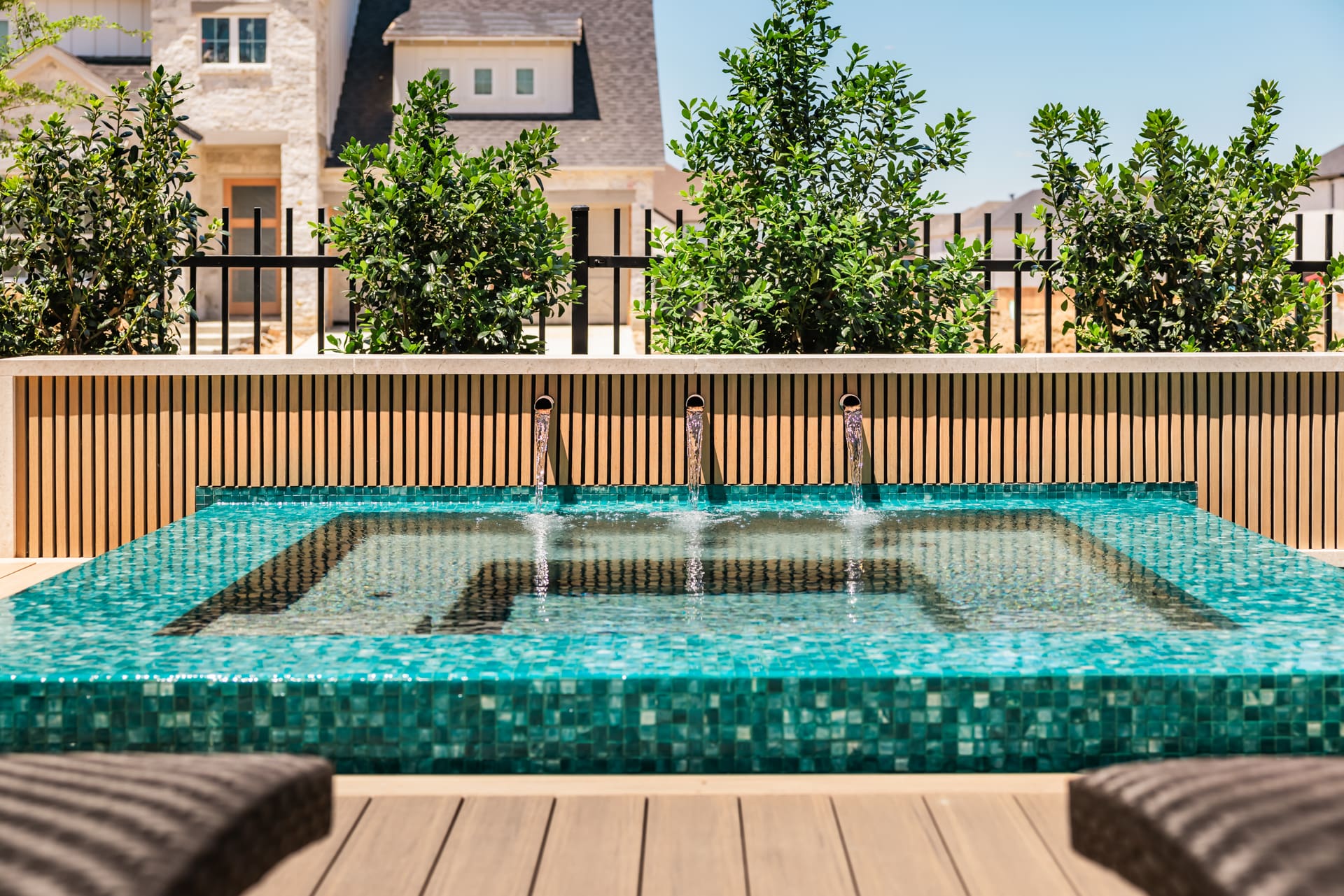 A luxurious outdoor hot tub with turquoise mosaic tiles and three spouts, set on a wooden deck. It is bordered by lush green bushes and set against a backdrop of a large, elegant house under a clear blue sky.