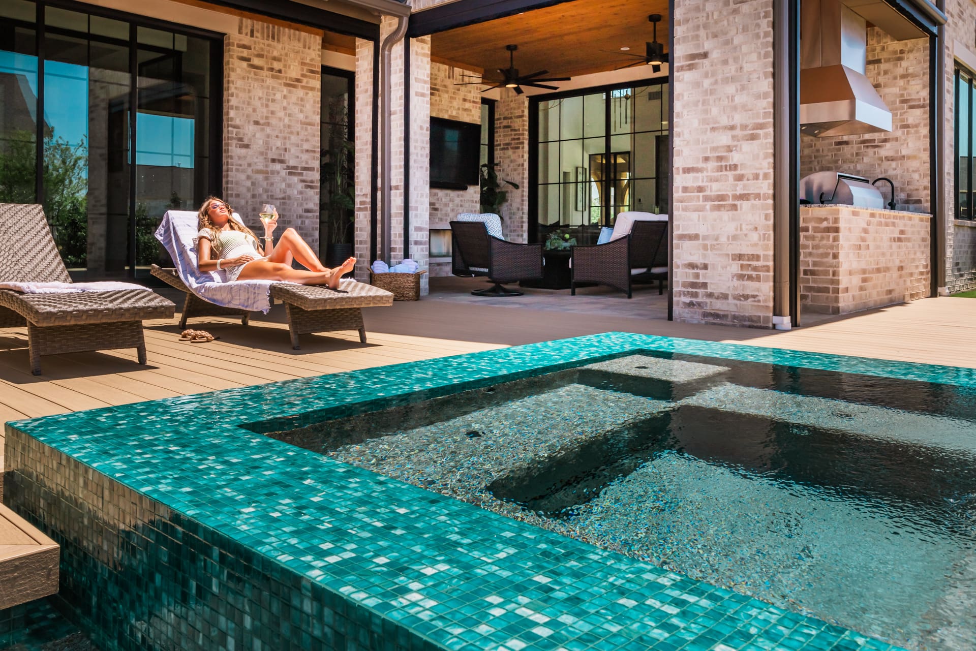 A woman relaxes on a lounge chair beside a modern outdoor pool with turquoise mosaic tiles. The spacious patio features comfortable seating and a built-in grill area, set against a backdrop of a brick house under a clear blue sky.