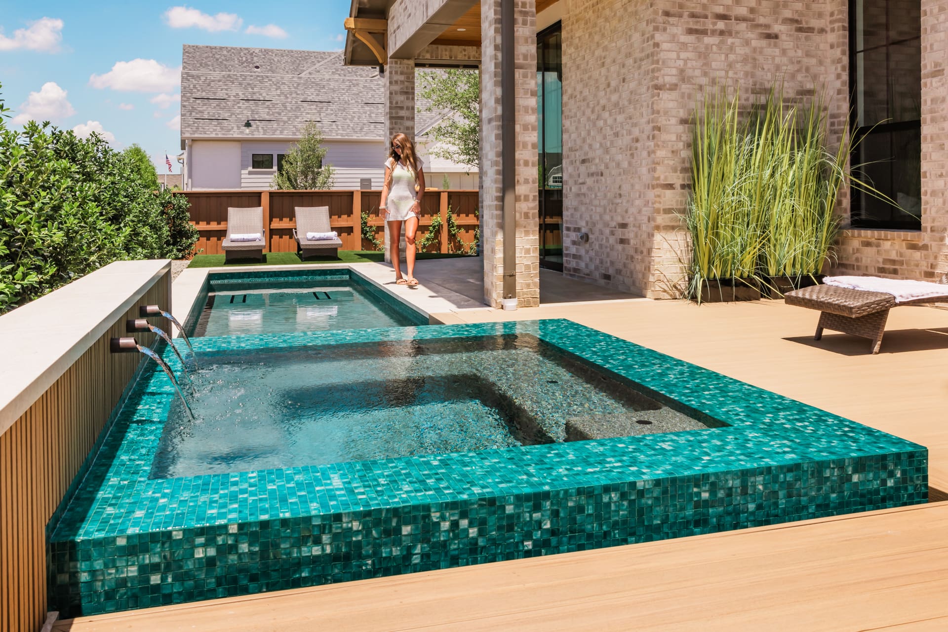 A modern backyard features a small, square, turquoise-tiled pool with water jets. A woman in a white dress stands near the pool, and lounge chairs are visible on the wooden deck. A light brick house and greenery surround the area.