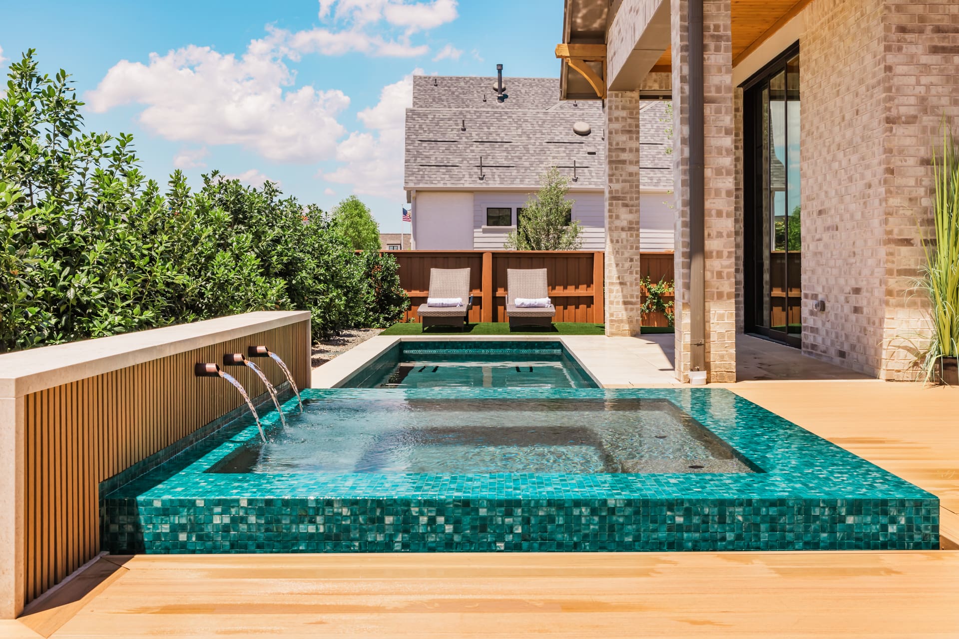 A modern backyard features a small rectangular turquoise pool with three spouts, surrounded by a wooden deck. Two lounge chairs rest on a grassy area. The setting is bordered by greenery and adjacent to a brick house under a partly cloudy sky.